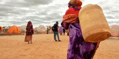 Yaşlı bir kadın, Somali'nin Doolow kentinde yaklaşık 3.000 yerinden edilmiş kişiye ev sahipliği yapan resmi olmayan yerleşim yerinde bidon taşıyor. Burada yaşayan insanların çoğu kuraklık nedeniyle yerinden edilmiş durumda. Fotoğraf: IOM 2022/Claudia Rosel
